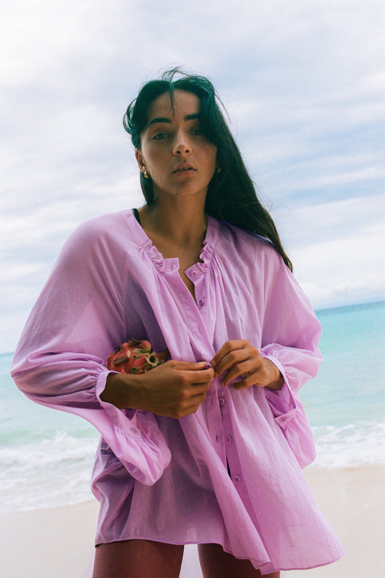 Thalia Cotton Voile Shirt in Orchid Pink - woman holding dragon fruit, gathered ruffle neckline and button-front, balloon sleeves, worn open as beach cover-up, ocean backdrop - Lilly Pilly Collection

