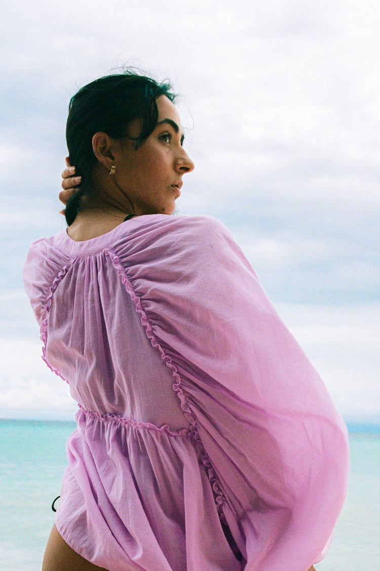 Thalia Cotton Voile Shirt in Orchid Pink - back view, gathered back yoke and balloon sleeves, hand in hair, worn open as beach cover-up, ocean backdrop - Lilly Pilly Collection
