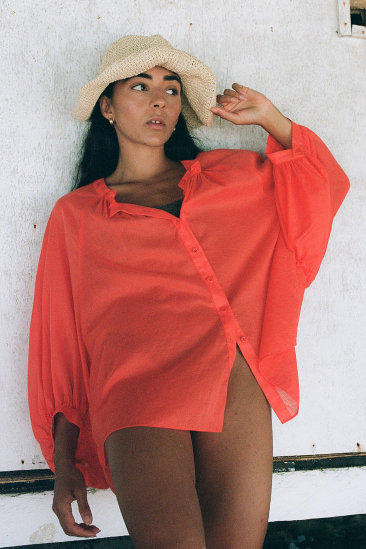 Thalia Cotton Voile Shirt in Spritz Orange - front view, gathered neckline and button-front, balloon sleeves, worn open as beach cover-up, straw hat, rustic white wall backdrop - Lilly Pilly Collection